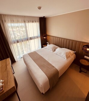 A hotel room with a large bed, a bedside table, a nightstand, and a view of the outdoors through sheer curtains.