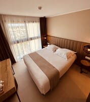 Habitación de hotel con una cama grande, una mesita de noche y vistas al exterior a través de cortinas translúcidas.