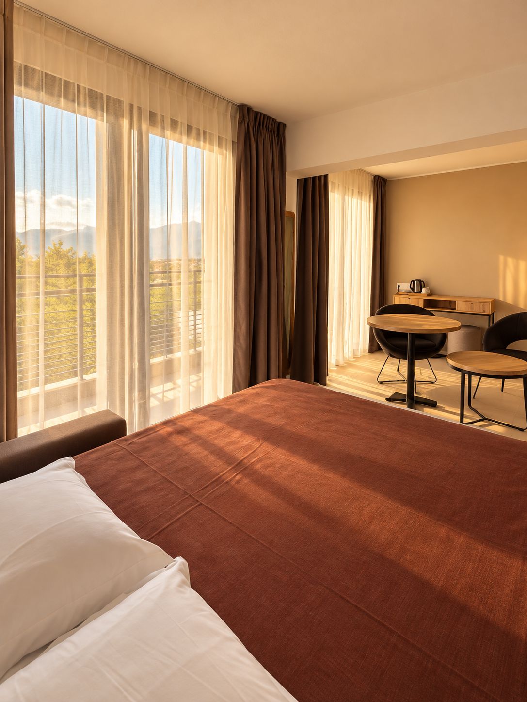 A hotel room with a large bed, a small round table, and a chair, a desk with a coffee maker, and a view of the outdoors through sheer curtains.