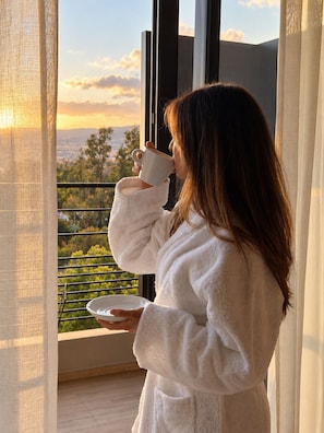 A person in a white bathrobe holding a cup and a plate, standing by a window with a view of greenery and a cityscape. - Hôtel Spa La Madrague (Lucciana)