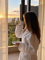Una persona con bata blanca sostiene una taza y un plato, de pie junto a una ventana con vista a zonas verdes y un paisaje urbano.