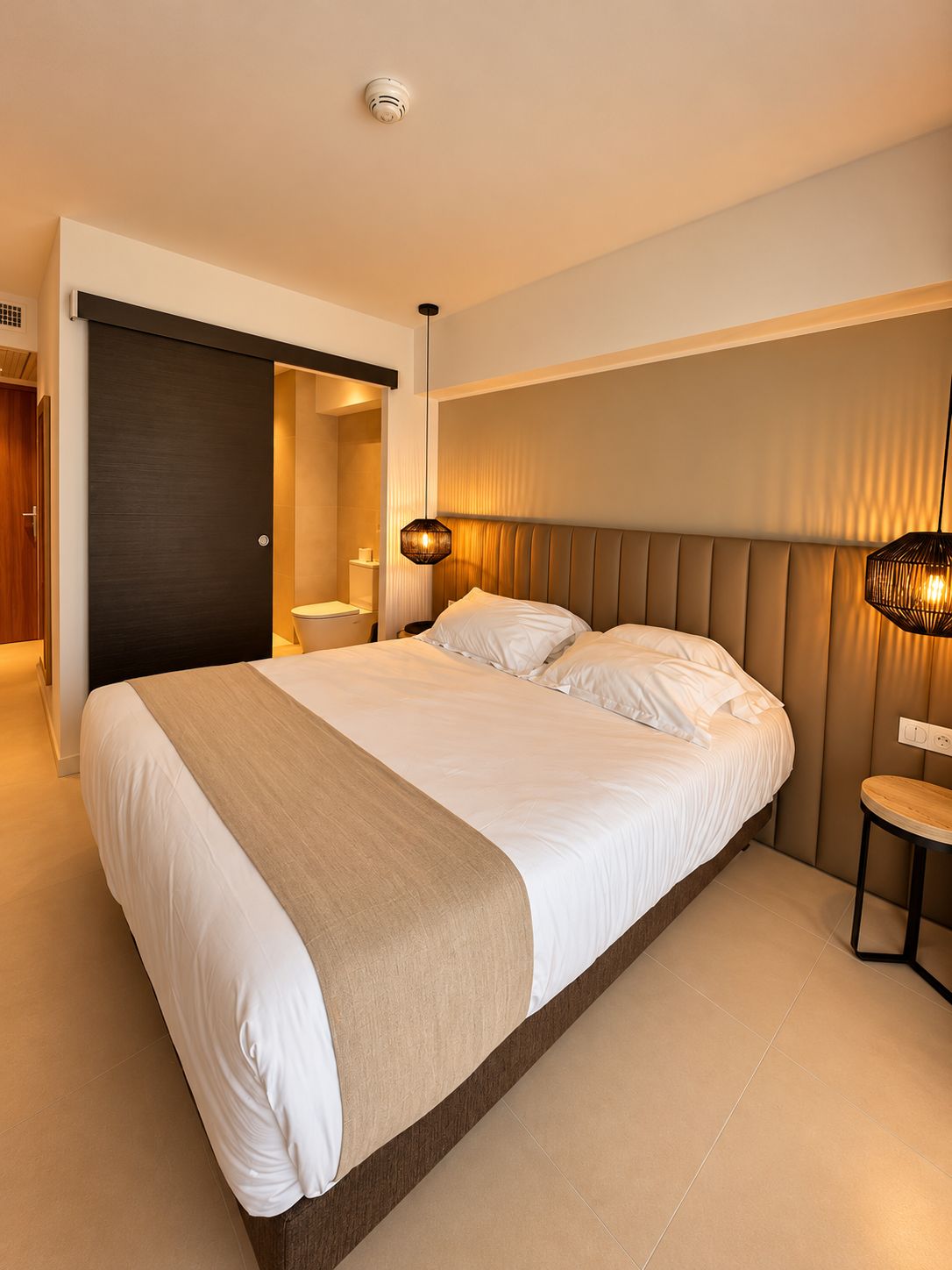 Une chambre d’hôtel moderne avec un grand lit, une table de chevet, un luminaire fixé au mur et une porte donnant sur une autre pièce.