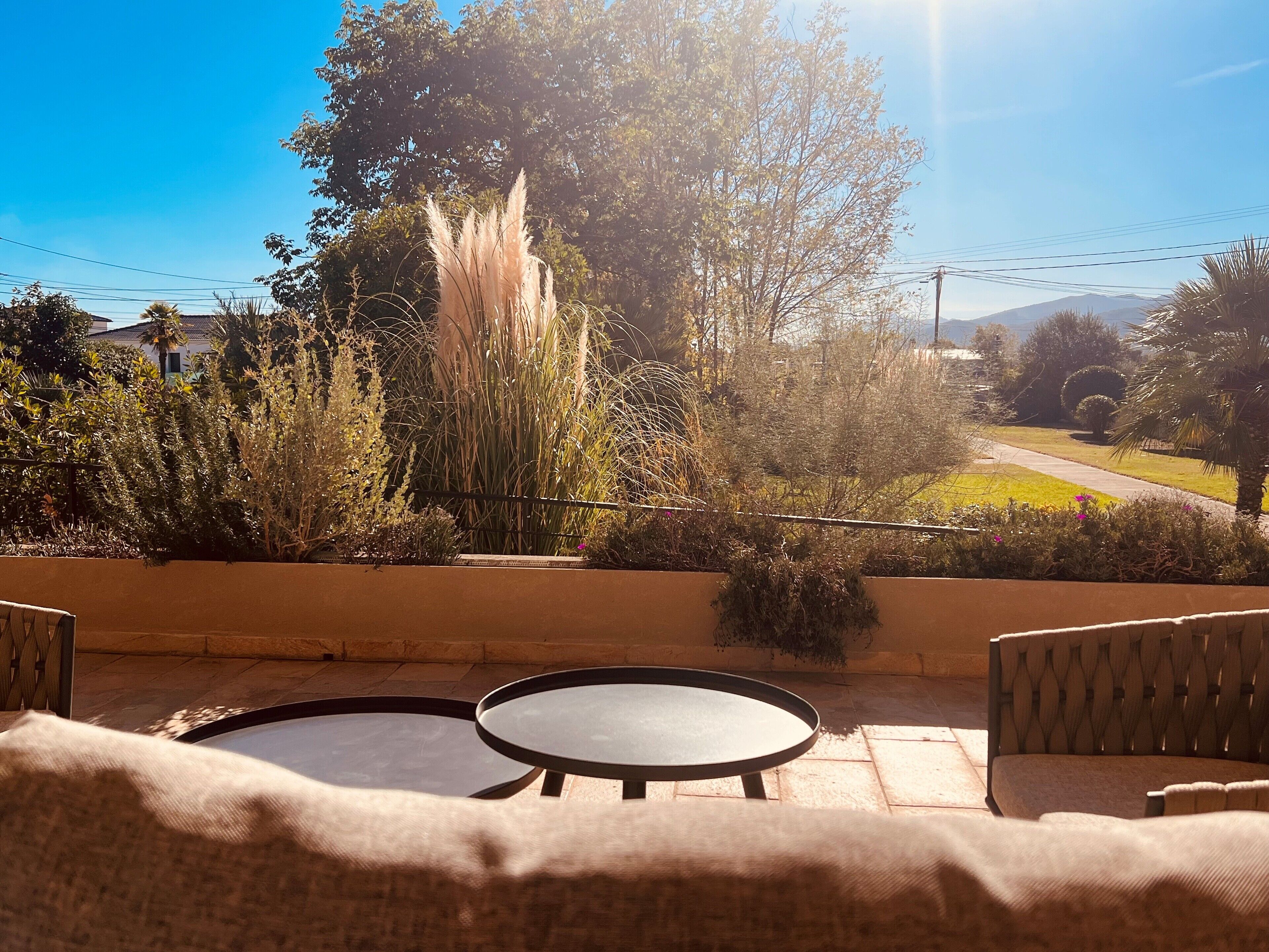 An outdoor seating area with two round tables and chairs, surrounded by a variety of plants and trees, with a clear view of the landscape in the background.
