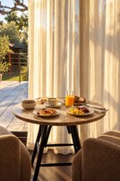 A round table with breakfast items and a glass of orange juice, set against a backdrop of curtains and a garden view.