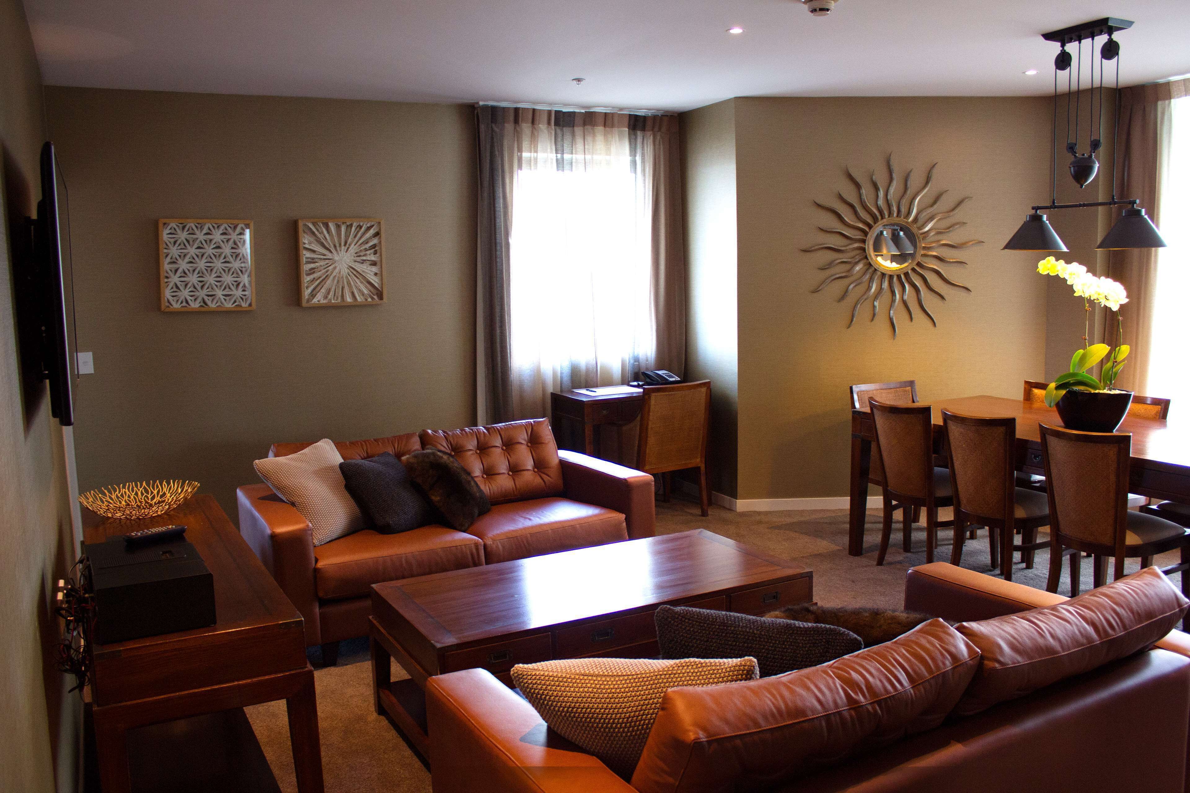 A living room with a sofa, coffee table, and dining area with chairs. There is a TV on the left and a decorative sun on the wall.