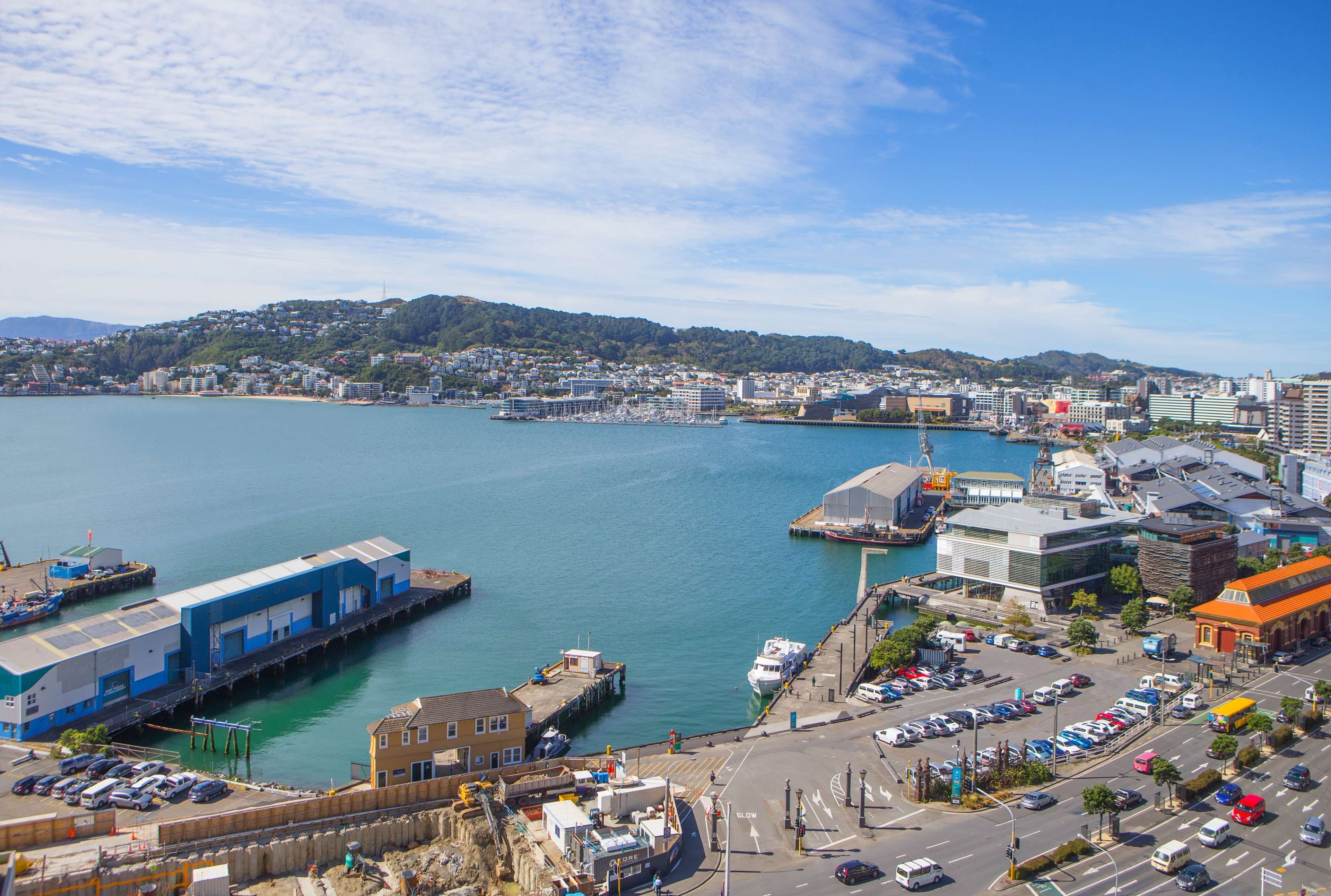 Un paisaje urbano costero con un puerto deportivo, edificios industriales y una calle con coches estacionados.