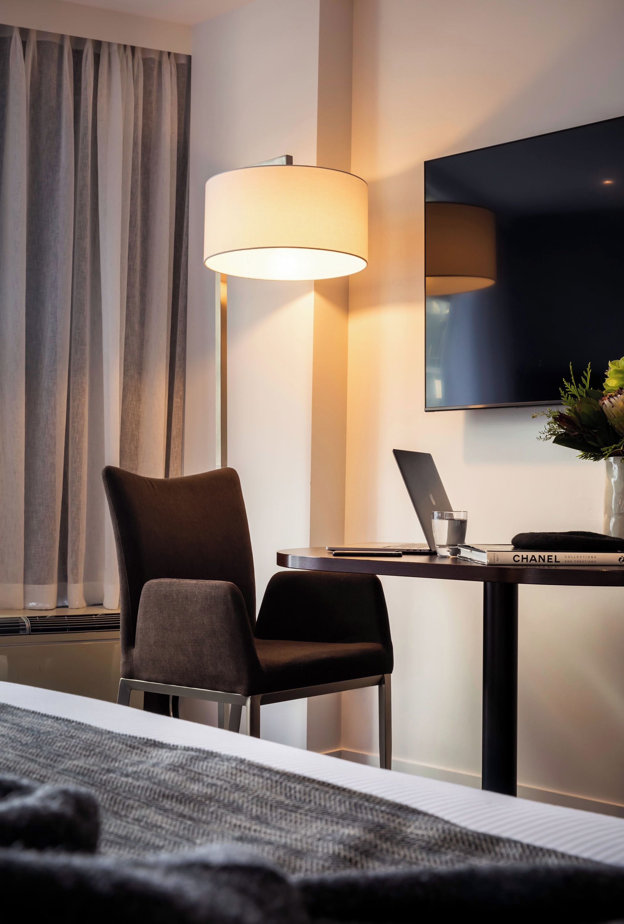 A hotel room with a bed, a desk with a laptop, a lamp, a TV, and a vase with flowers.