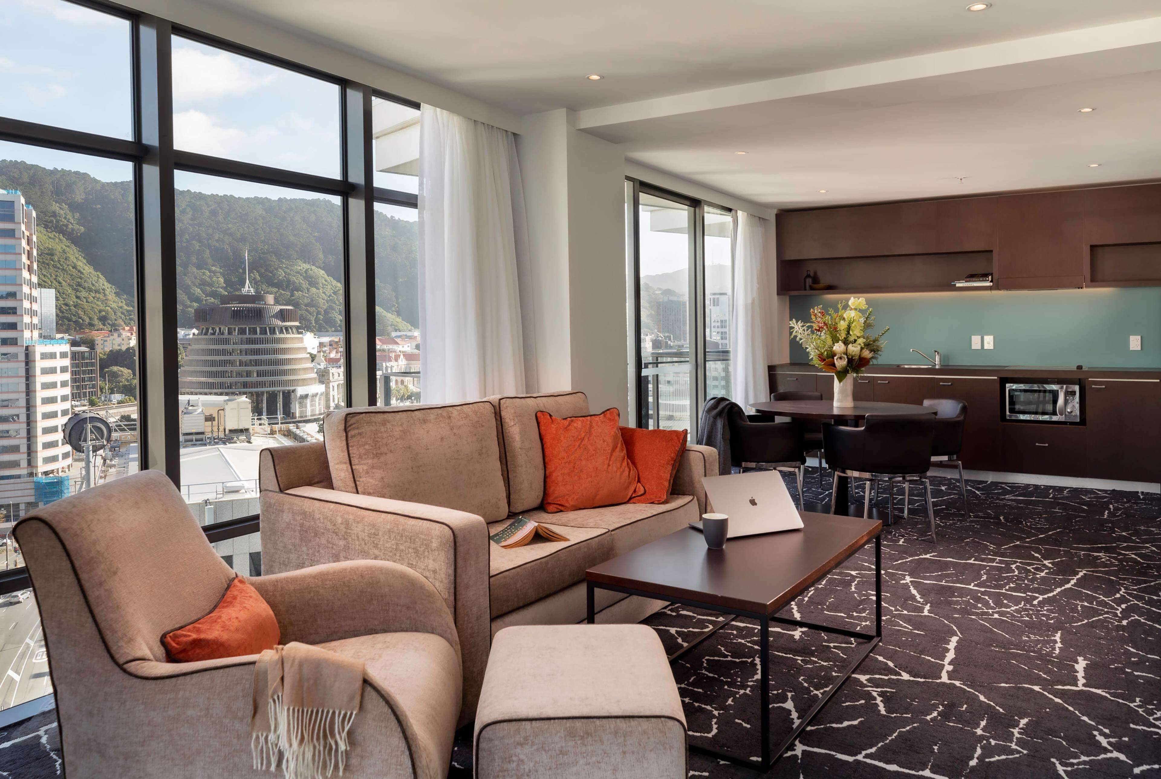 A modern hotel room with a large window offering a city view, a sofa set, a dining table, and a kitchen area.
