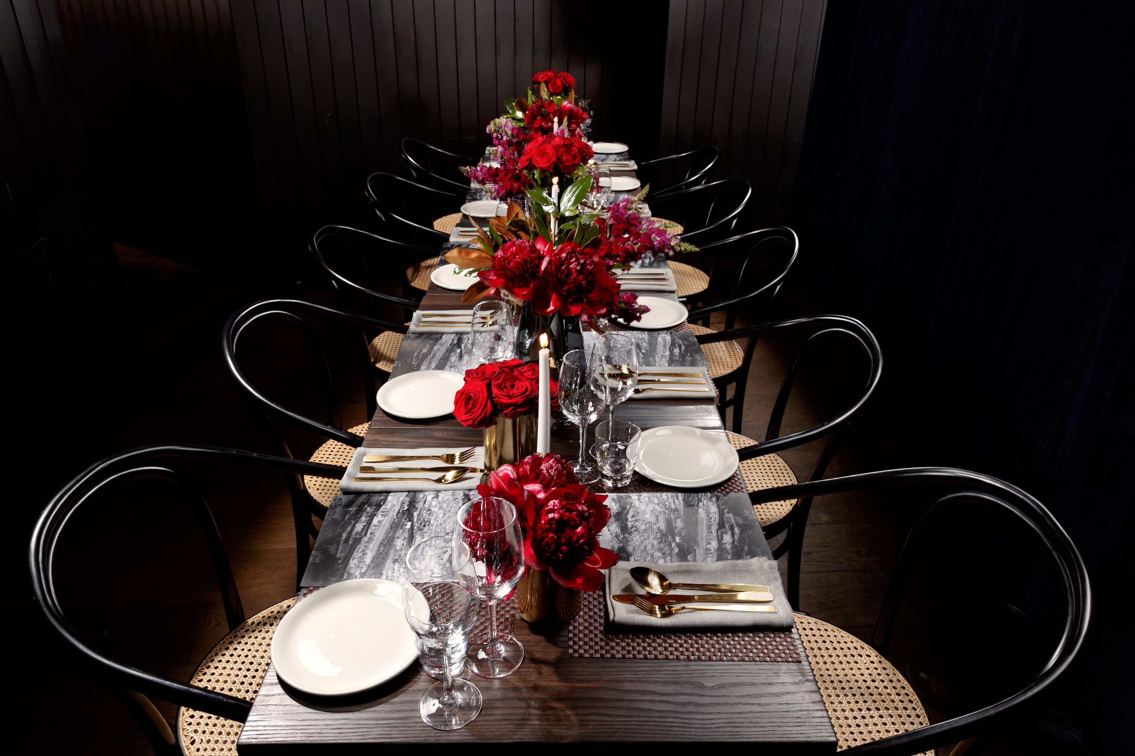 A long dining table set with red flowers, clear glasses, and gold cutlery.