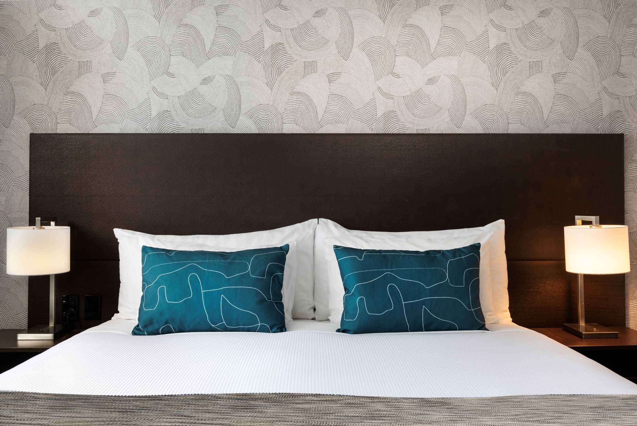 A neatly made bed with white bedding and two teal pillows, flanked by two bedside tables with lamps, against a patterned wallpaper backdrop.