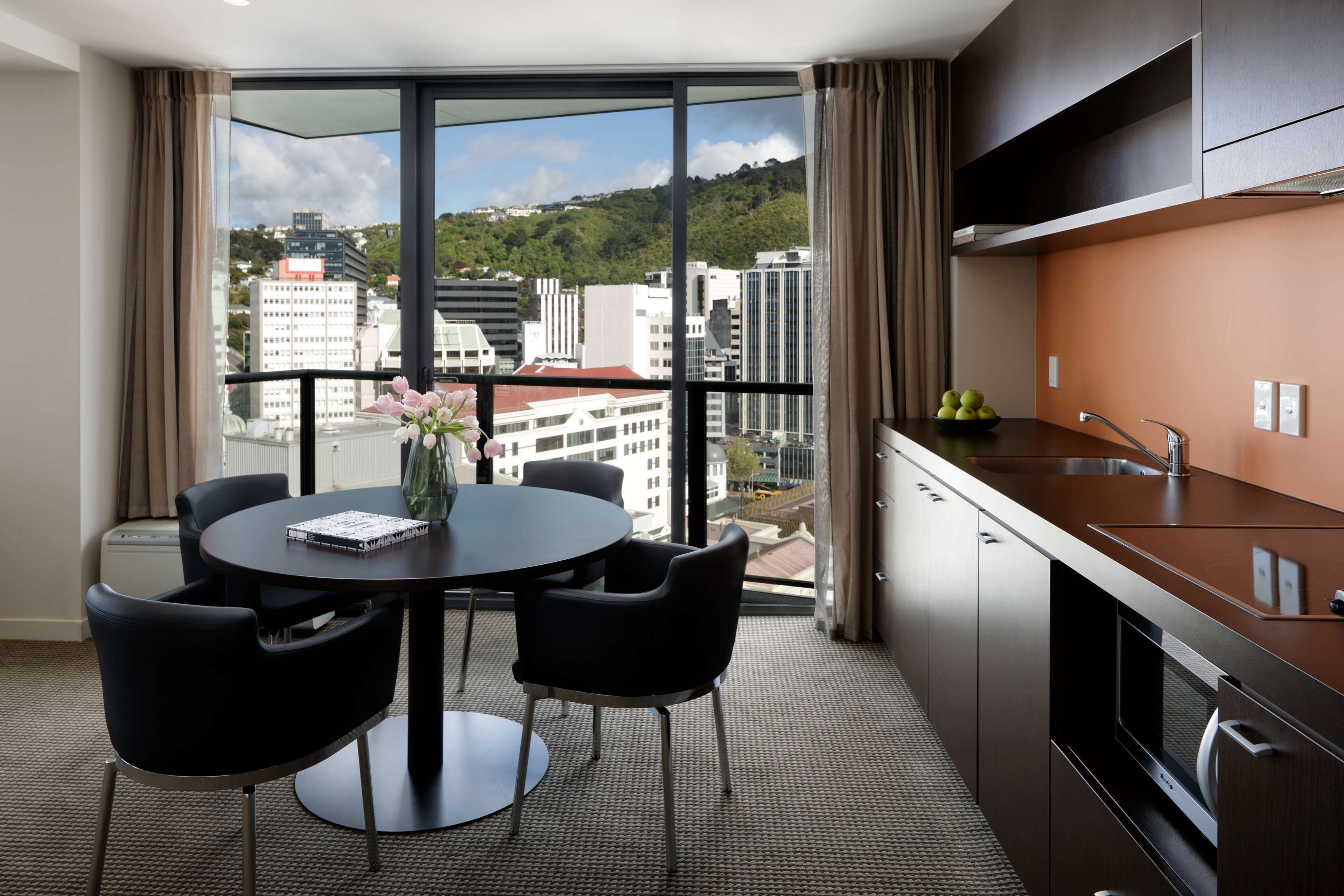 A modern hotel room with a dining area, a large window with a city view, a kitchen with a sink, and a flat-screen TV.
