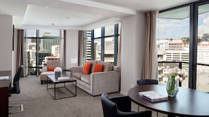 A modern hotel room with a large window offering a city view, a comfortable sofa, a dining table, and a workspace with a desk and chair. - Rydges Wellington (Wellington)