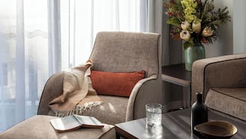 A modern living room with a comfortable armchair, a small table with a book, a glass of water, a vase with flowers, and a view of a cityscape through sheer curtains.