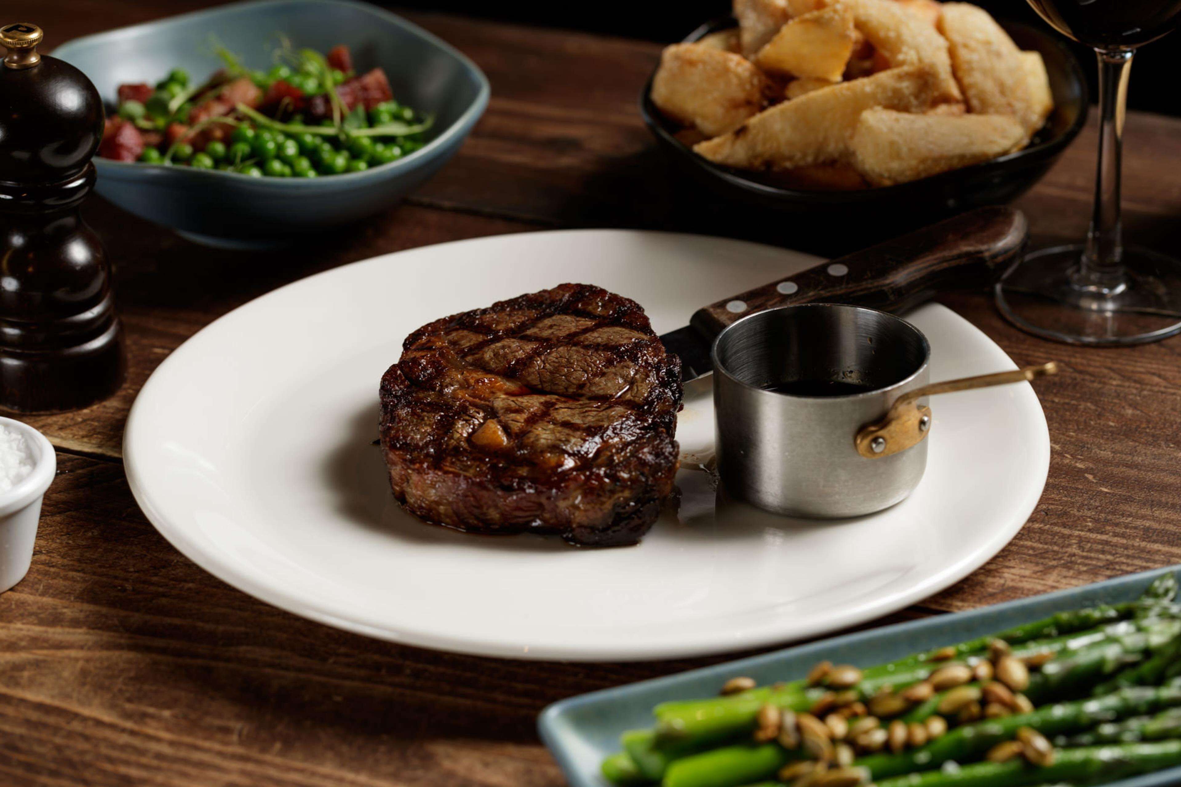 Bife num prato com um pequeno recipiente de molho, aspargos e uma salada numa mesa de madeira.