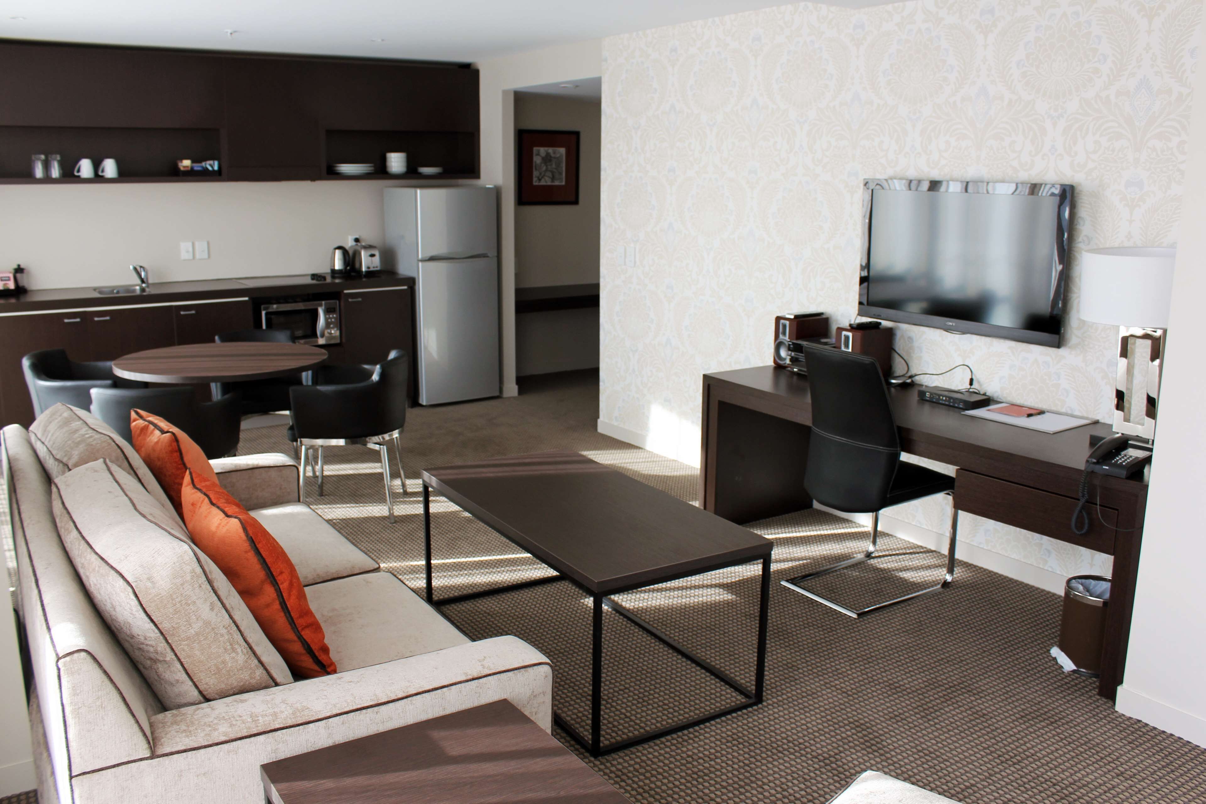 Habitación de hotel moderna con sofá, mesa de centro, mesa de comedor, sillas, zona de cocina y televisor.