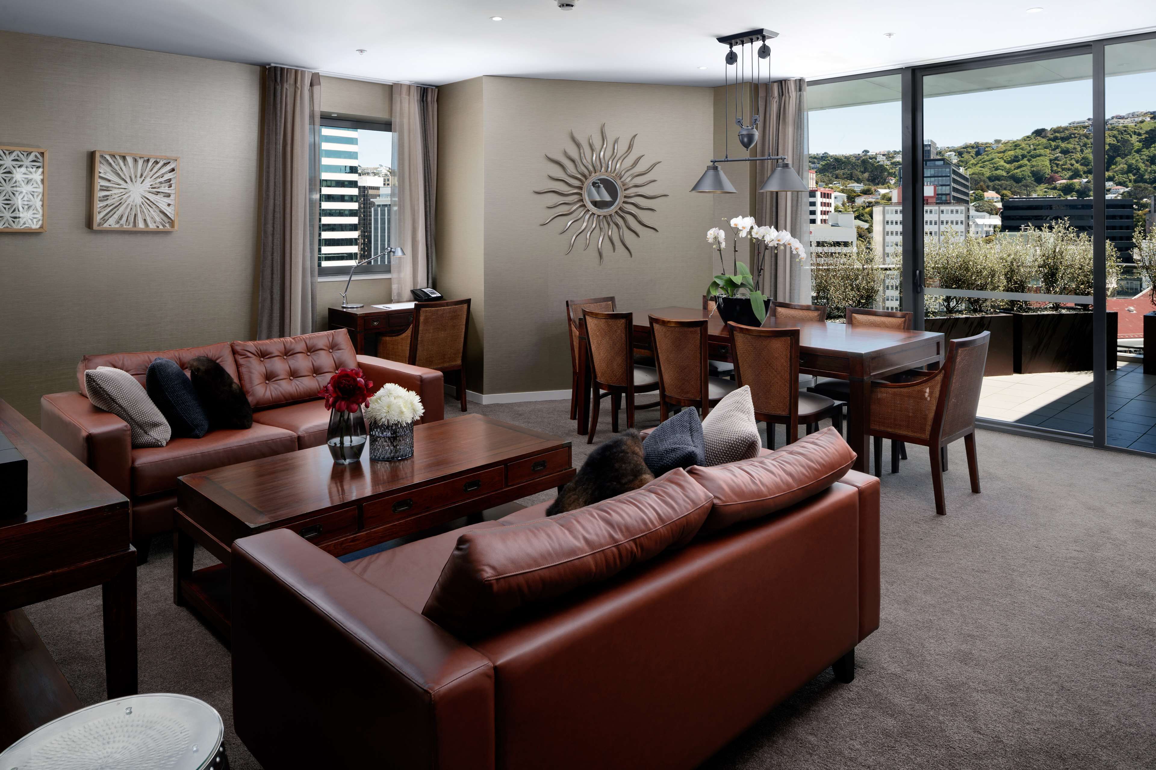 A modern living room with a brown leather sofa, a dining table with chairs, and a large window offering a city view.