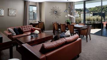 A modern living room with a brown leather sofa, a dining table with chairs, and a large window offering a city view.