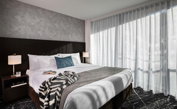 A hotel room with a large bed, two bedside lamps, a nightstand with a glass of water and a book, and sheer curtains allowing natural light.