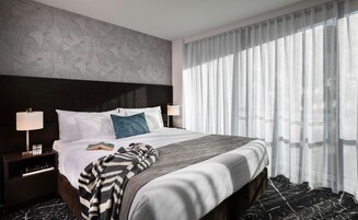 Camera d'albergo con un letto grande, due lampade ai comodini, un comodino con un bicchiere d'acqua e un libro, e tende leggere che lasciano filtrare la luce naturale.
