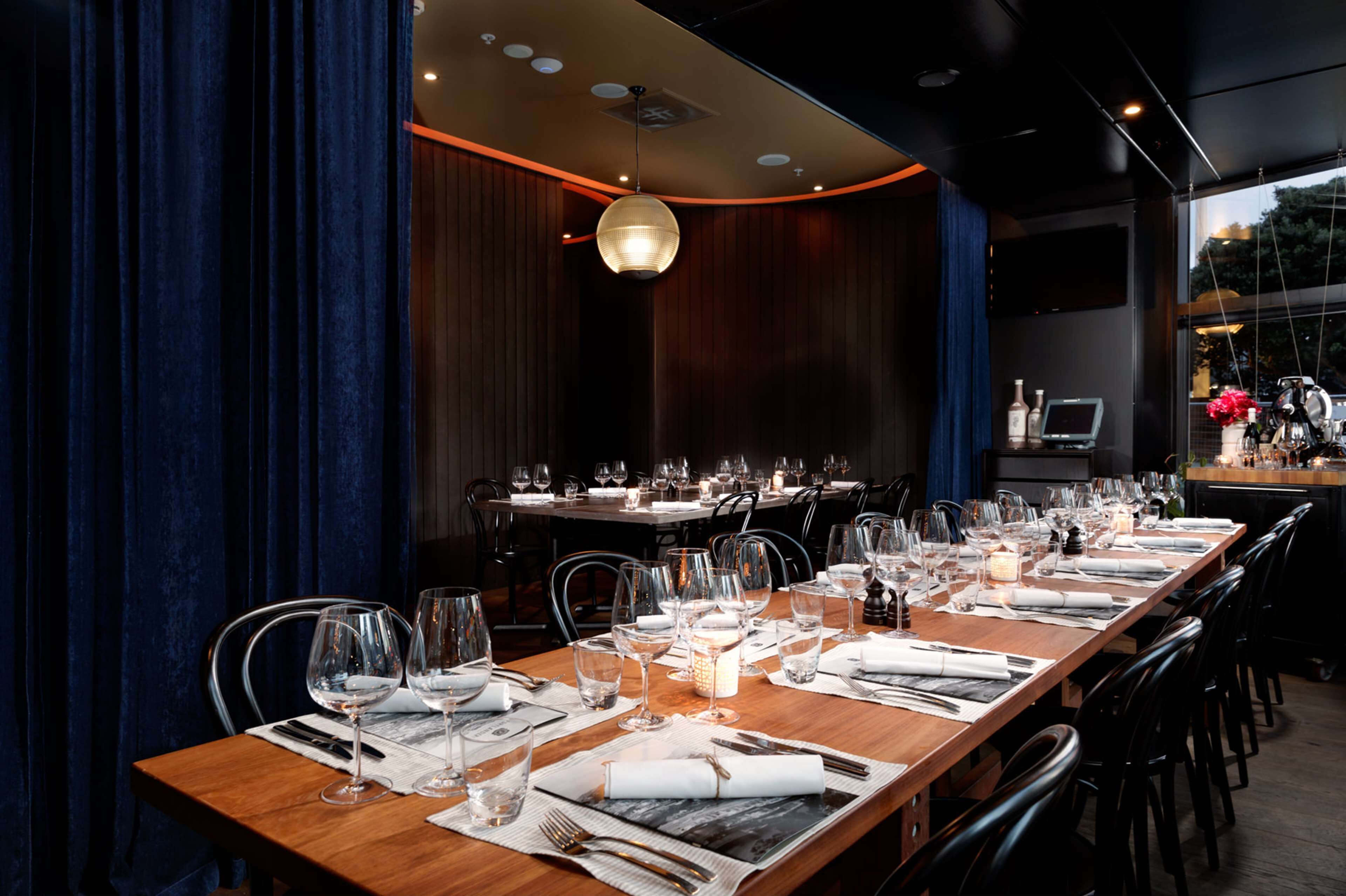 Um restaurante com uma longa mesa de madeira posta para refeição, rodeada por cadeiras e uma luminária pendente moderna.