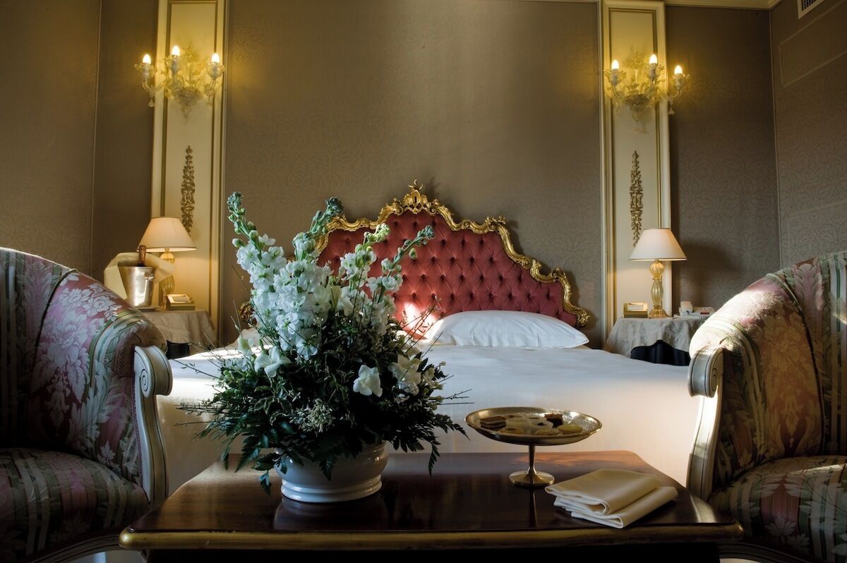 A hotel room with a tufted red headboard, floral arrangements, two lamps, and a bedside table with a tray.