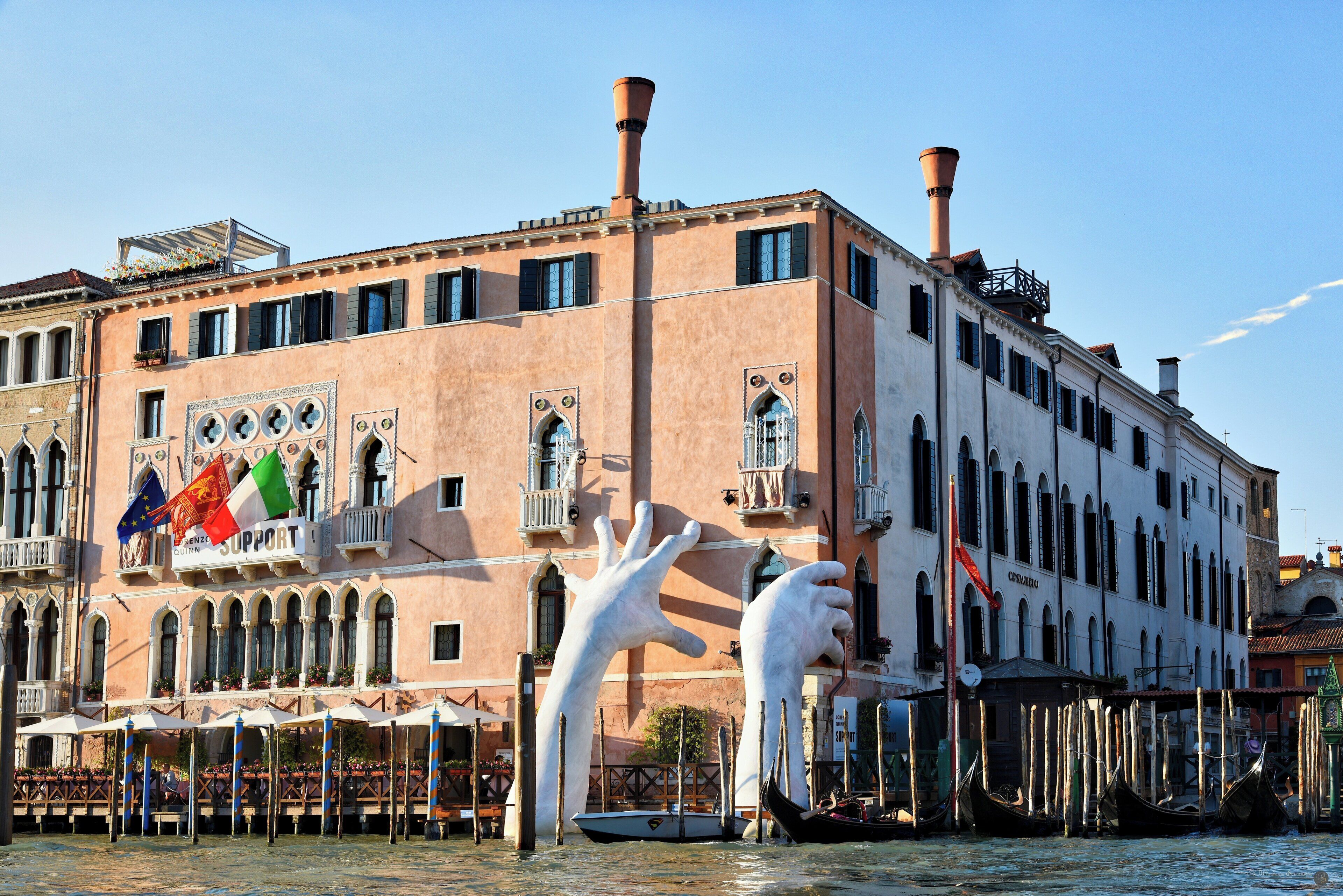 A historic building with a large sculpture in front, a canal with gondolas, and flags.