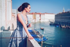 A woman in a patterned dress leaning on a balcony railing overlooking a canal with boats and buildings.