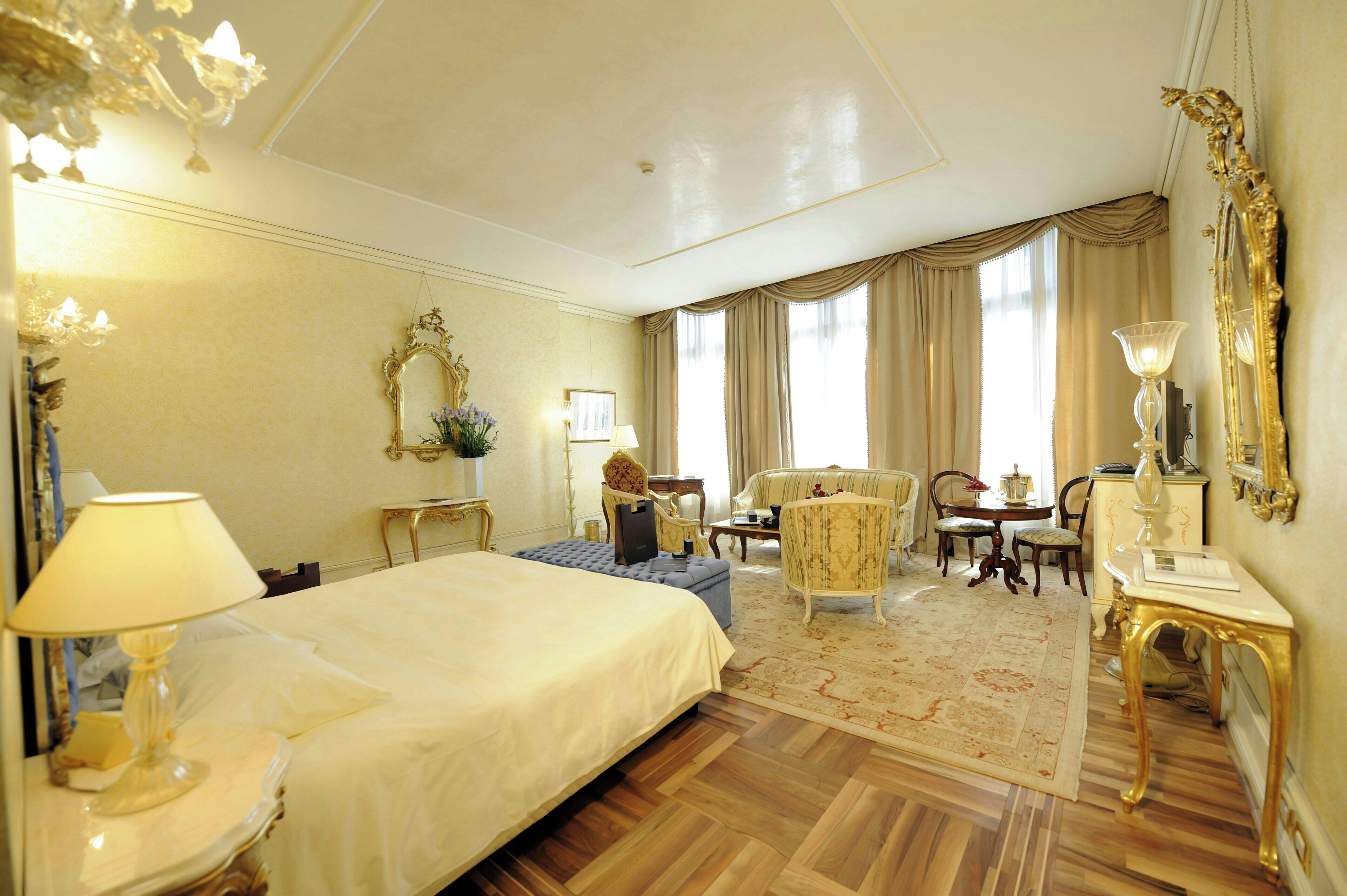 Une chambre d’hôtel dotée d’un grand lit, d’un coin salon avec un canapé et des fauteuils, d’une table à manger et d’un lustre.