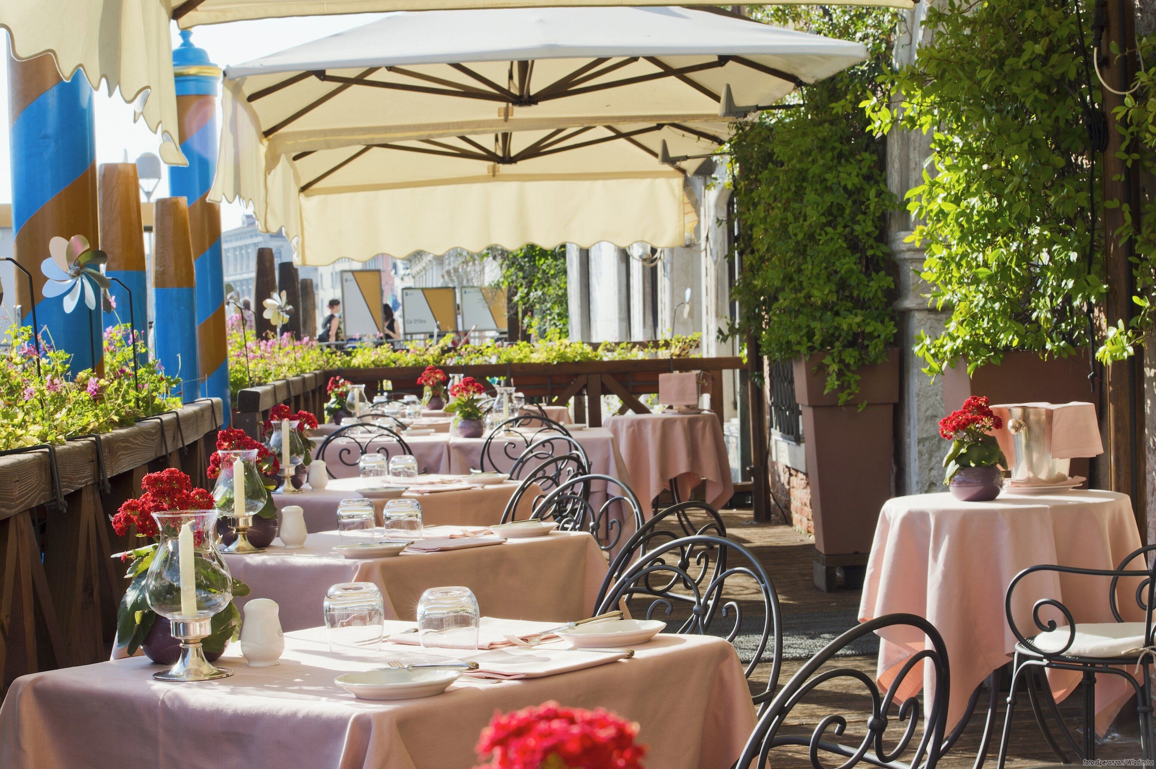 An outdoor dining area with tables set for two, covered by umbrellas, and surrounded by greenery.