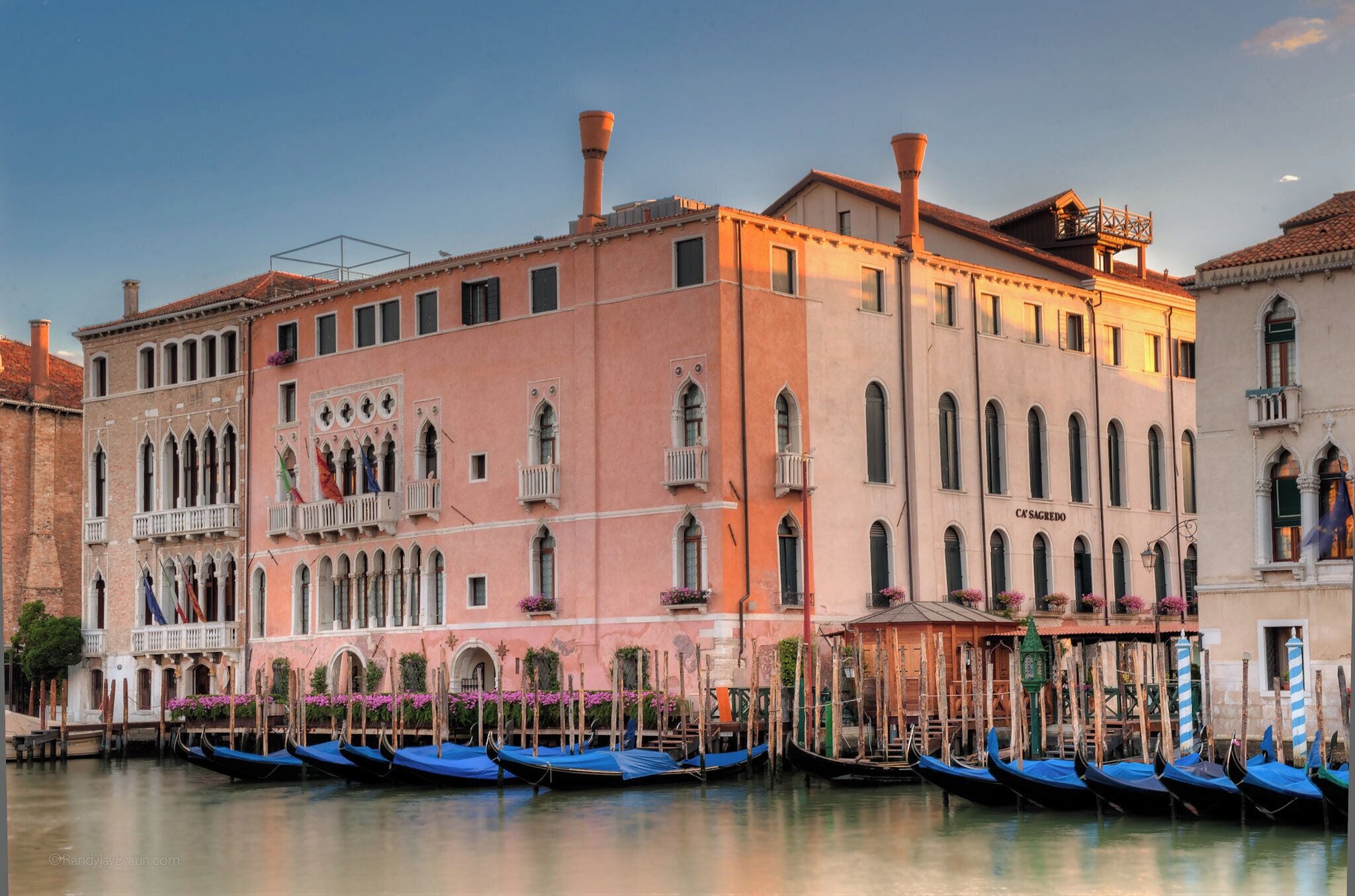 A historic building with multiple windows and balconies, a sign reading "C'EST S'AGREDO", and several gondolas docked in the water.
