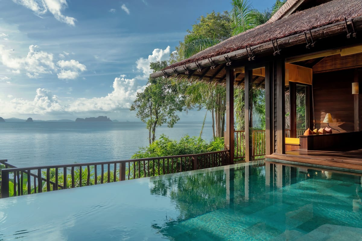 A clear view of an infinity pool with a wooden deck, a thatched roof structure, and a view of the sea.