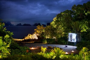 A beachside setting with a large screen displaying a scenic view, surrounded by lush greenery and a calm sea under a dark sky.