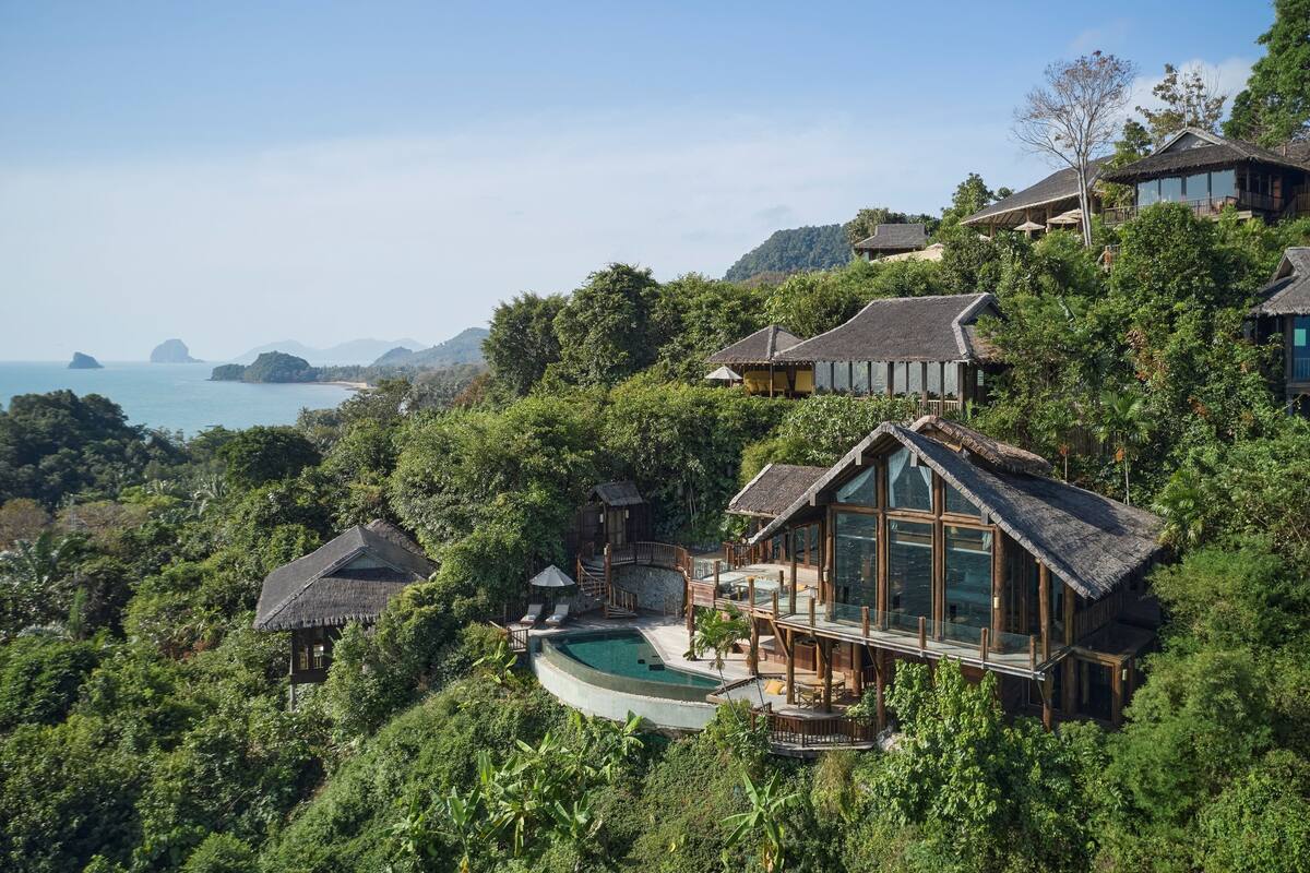 A hillside villa with a swimming pool, wooden deck, and multiple buildings surrounded by lush greenery and overlooking a coastal area.