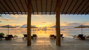 Un espacio de estar al aire libre cubierto con sillones y sombrillas, con vista a una masa de agua tranquila e islas distantes al atardecer.