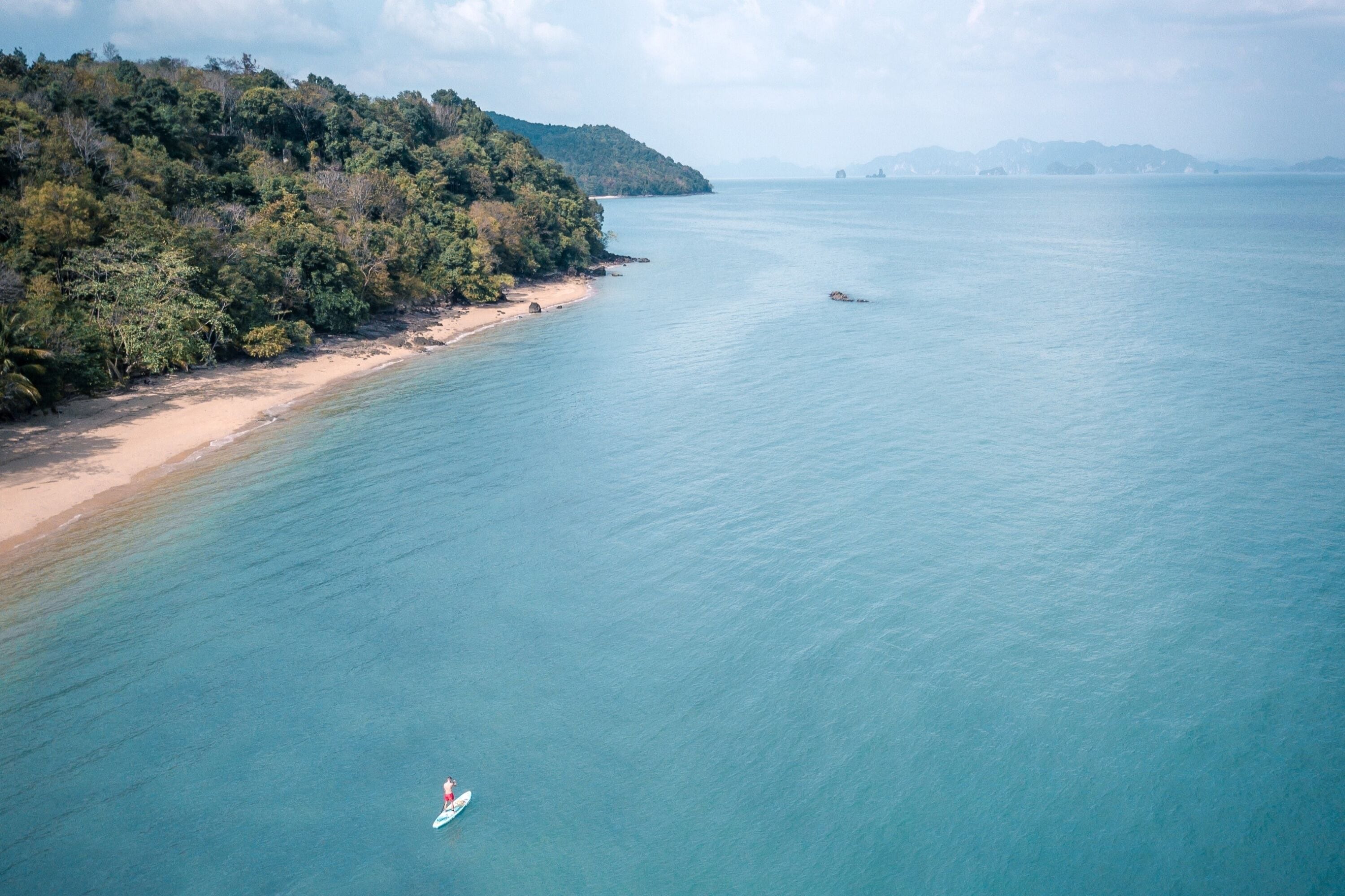 An aerial view of a beach with a clear sky, calm sea, and a person on a surfboard.