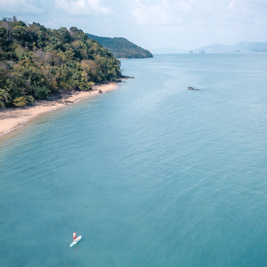 An aerial view of a beach with a clear sky, calm sea, and a person on a surfboard.