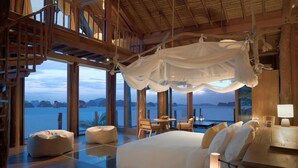 Un dormitorio amplio con una cama grande, un área de descanso con una mesa y sillas, y vista al mar a través de ventanas grandes.