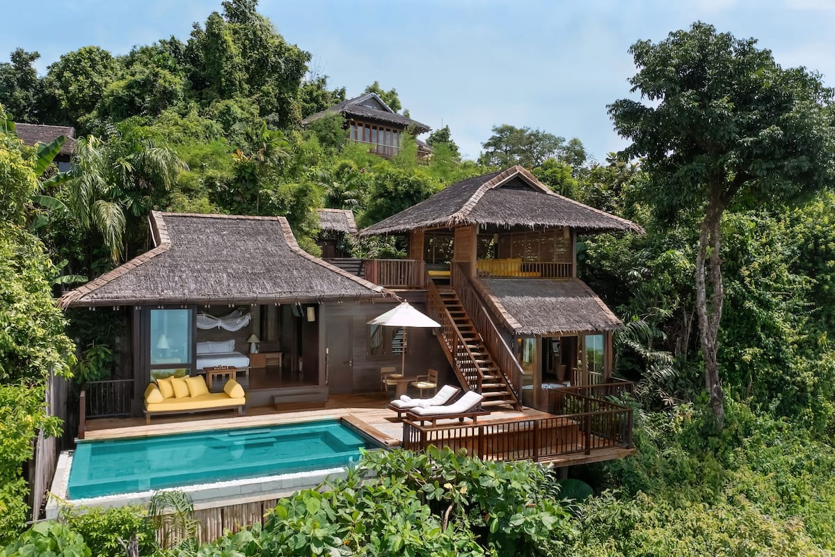 A resort with a pool, wooden deck, and thatched-roof buildings surrounded by lush greenery.