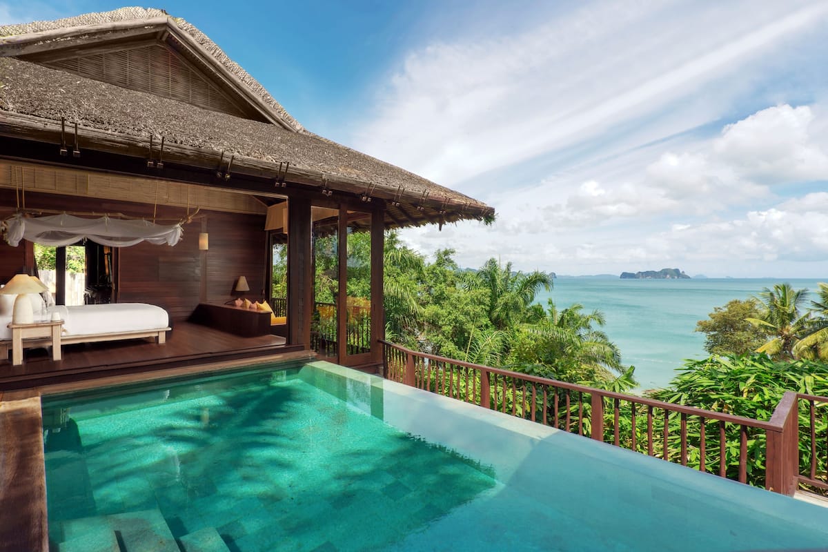 A poolside area with a wooden deck, a lounge chair, and a view of the ocean and palm trees.