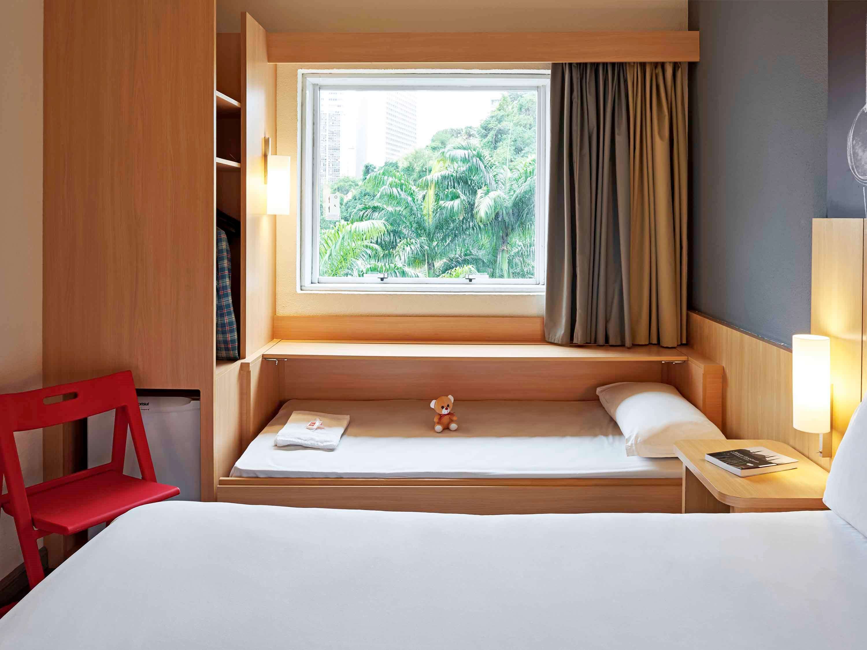 A hotel room with a bed, a small window with a view of greenery, a red chair, and a nightstand with a lamp.