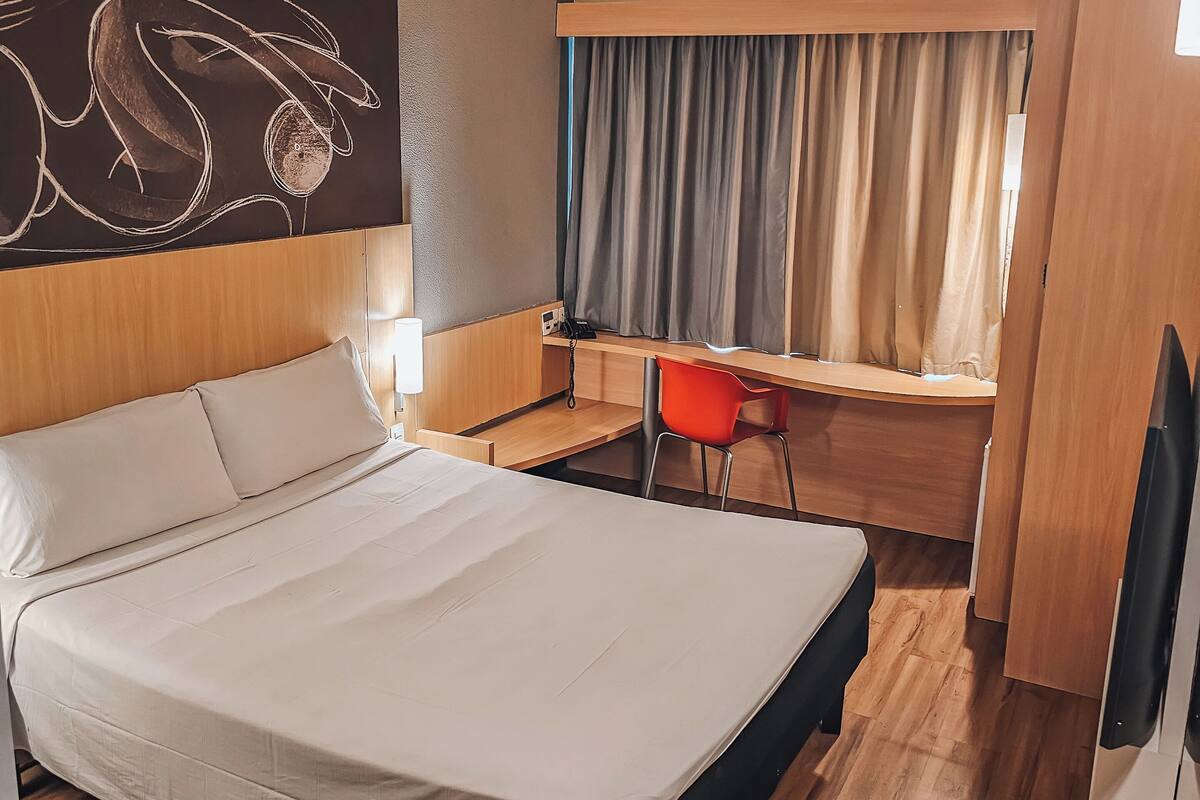 A hotel room with a large bed, a desk, a chair, and a window with curtains.