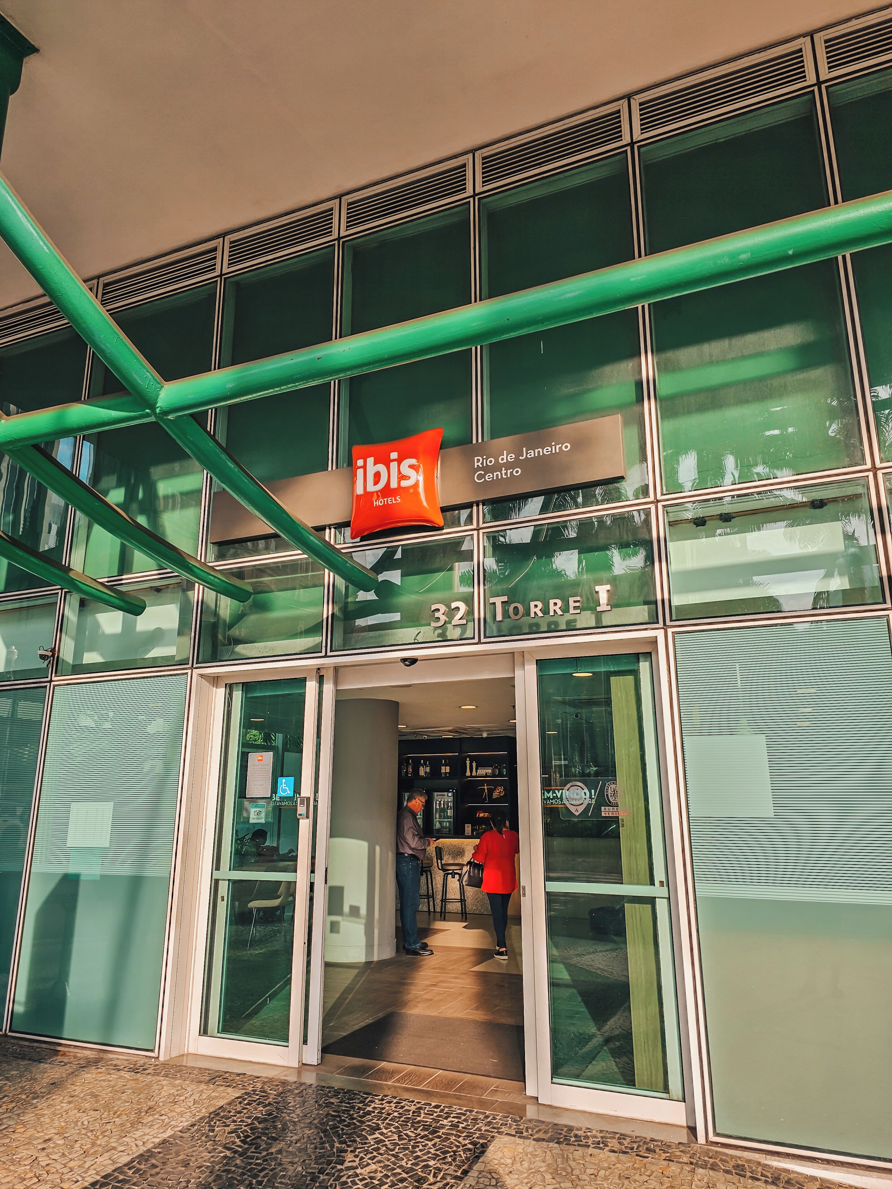 The exterior of the ibis Rio de Janeiro Centro hotel, featuring a modern glass facade, a prominent red and white sign, and a visible entrance with a tiled floor.