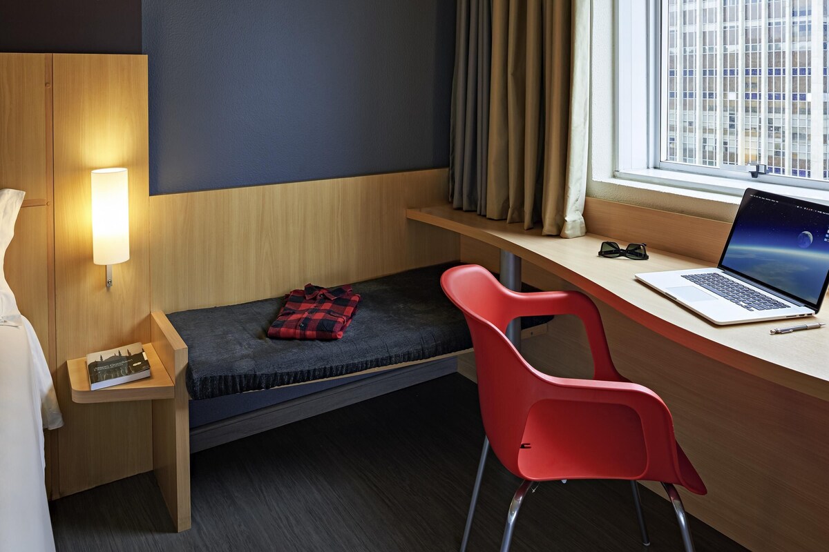 A compact hotel room with a single bed, a desk with a laptop, a red chair, and a window with curtains.