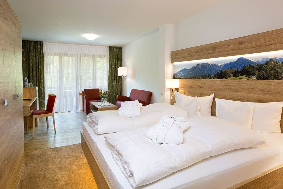 A hotel room with a large bed, two chairs, a desk, and a view of mountains.