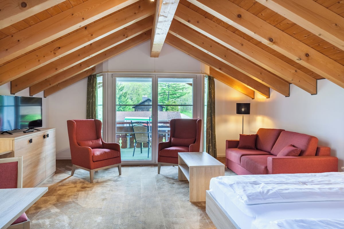 A cozy room with a sloped ceiling, a bed, a red sofa, a red armchair, a wooden table, and a TV on a wooden cabinet.
