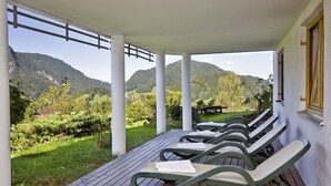 Een overdekt terras met houten banken, uitzicht op een bergachtig landschap en een wit gebouw met een raam.