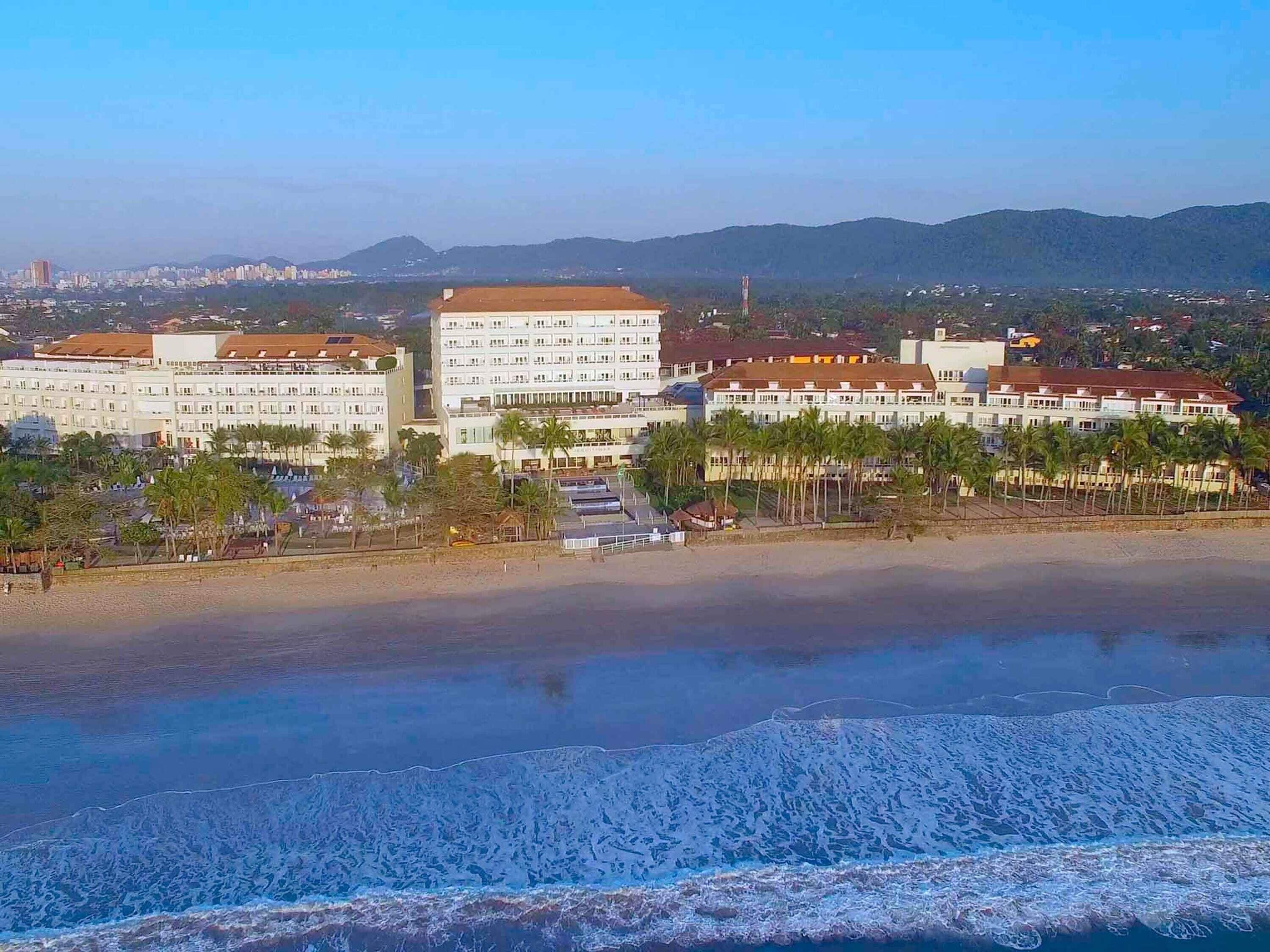 A coastal hotel with multiple buildings, a beach, and mountains in the background.