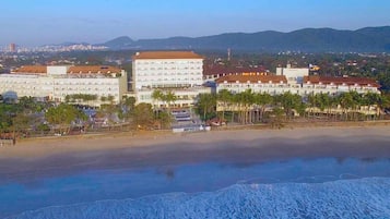 A coastal hotel with multiple buildings, a beach, and mountains in the background.