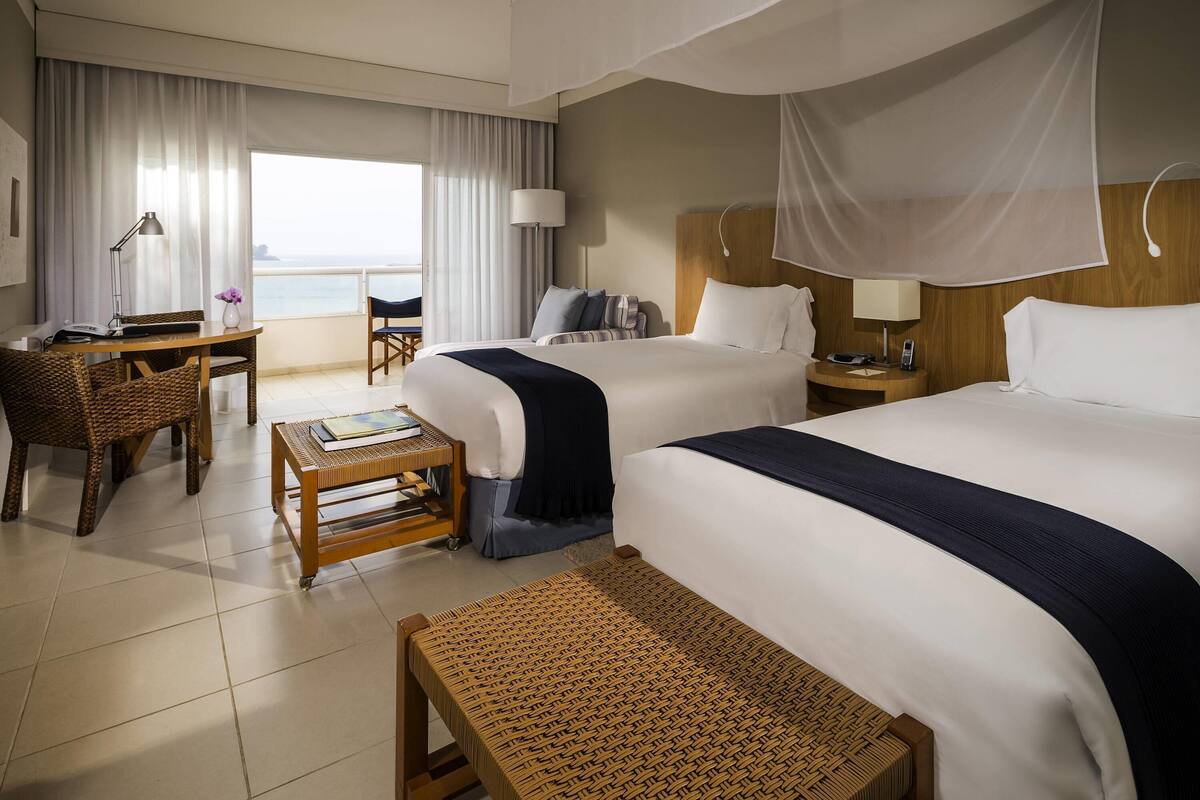 A hotel room with two beds, a desk, a chair, a small table, and a view of the sea.