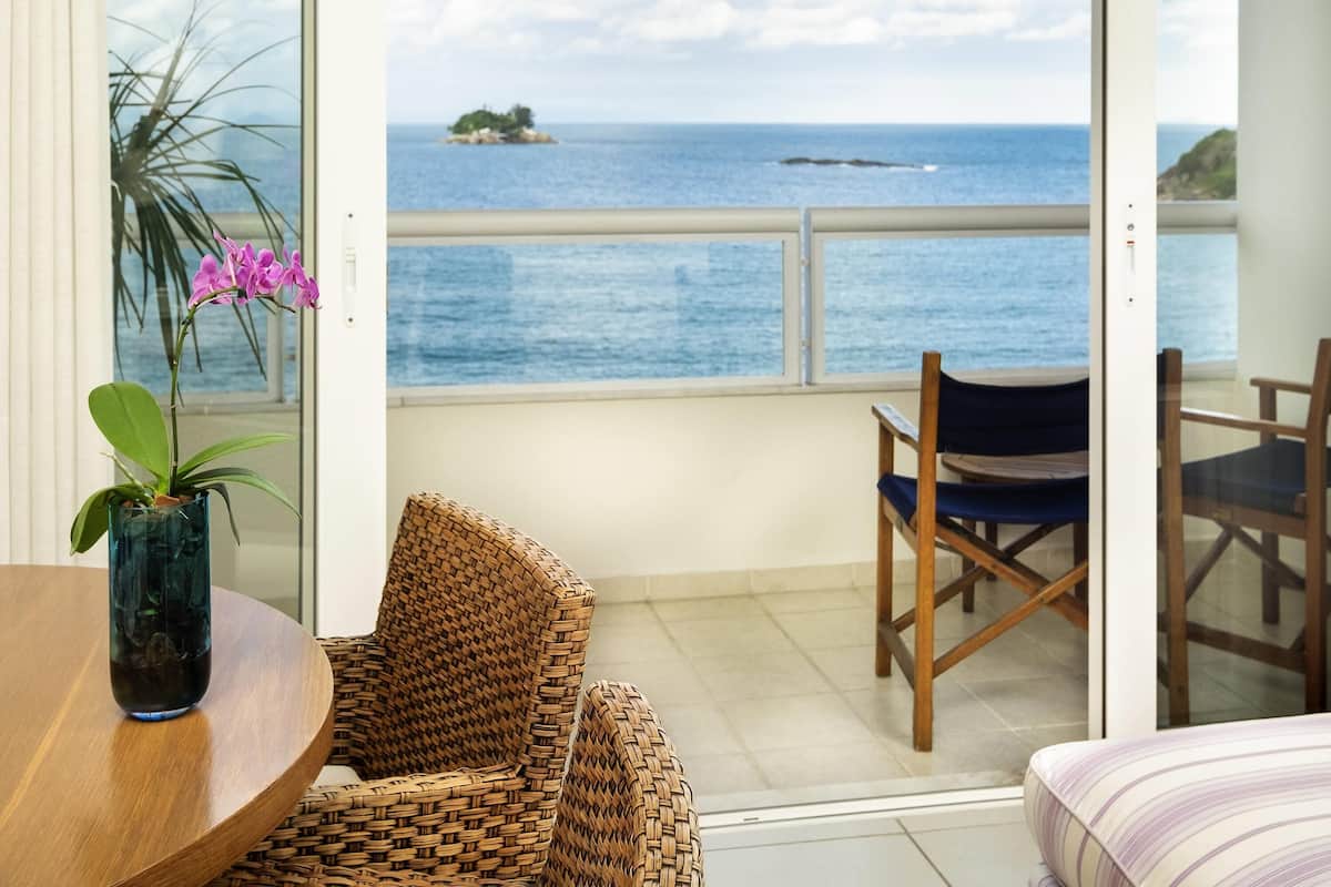 A balcony with a wicker chair, a small table with a vase of flowers, and a view of the sea.